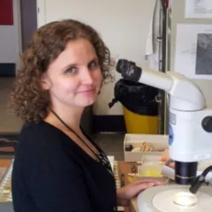 A light-skinned woman with curly hair sits in front of a microscope